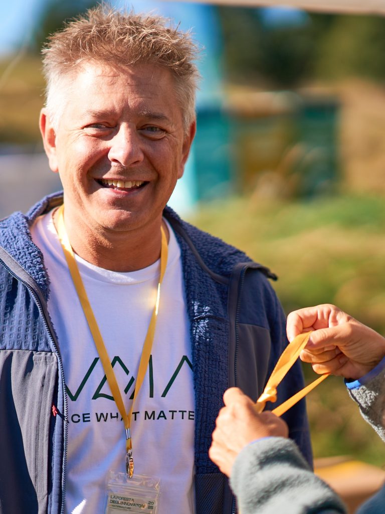 Alessandro Dietre, Co-founder & COO di VAIA Ritratto di Alessandro Dietre, co-fondatore e COO di VAIA, sorridente durante un evento all’aperto. Indossa un badge giallo e una t-shirt con il logo VAIA.