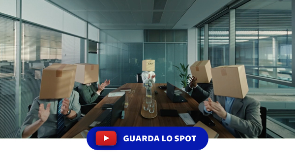 Pulsante "Guarda lo spot" e Frame dello spot GLS che mostra una riunione in ufficio con persone sedute attorno a un tavolo, ciascuna con una scatola di cartone in testa. La scena, surreale e ironica, rappresenta il concept creativo della campagna Out of Home “Ci pensiamo noi”.