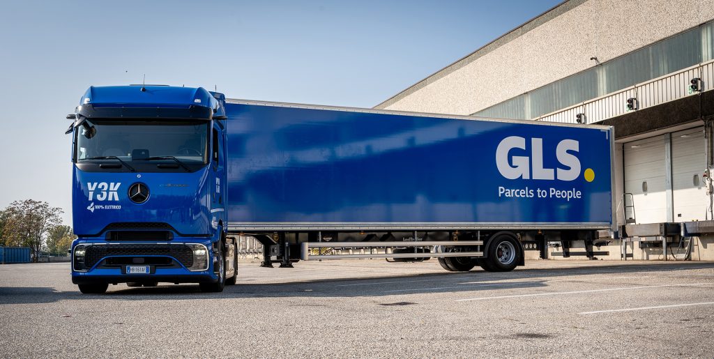 Un truck elettrico blu parcheggiato fuori da un magazzino, con il logo GLS e lo slogan “Parcels to People” sul rimorchio.